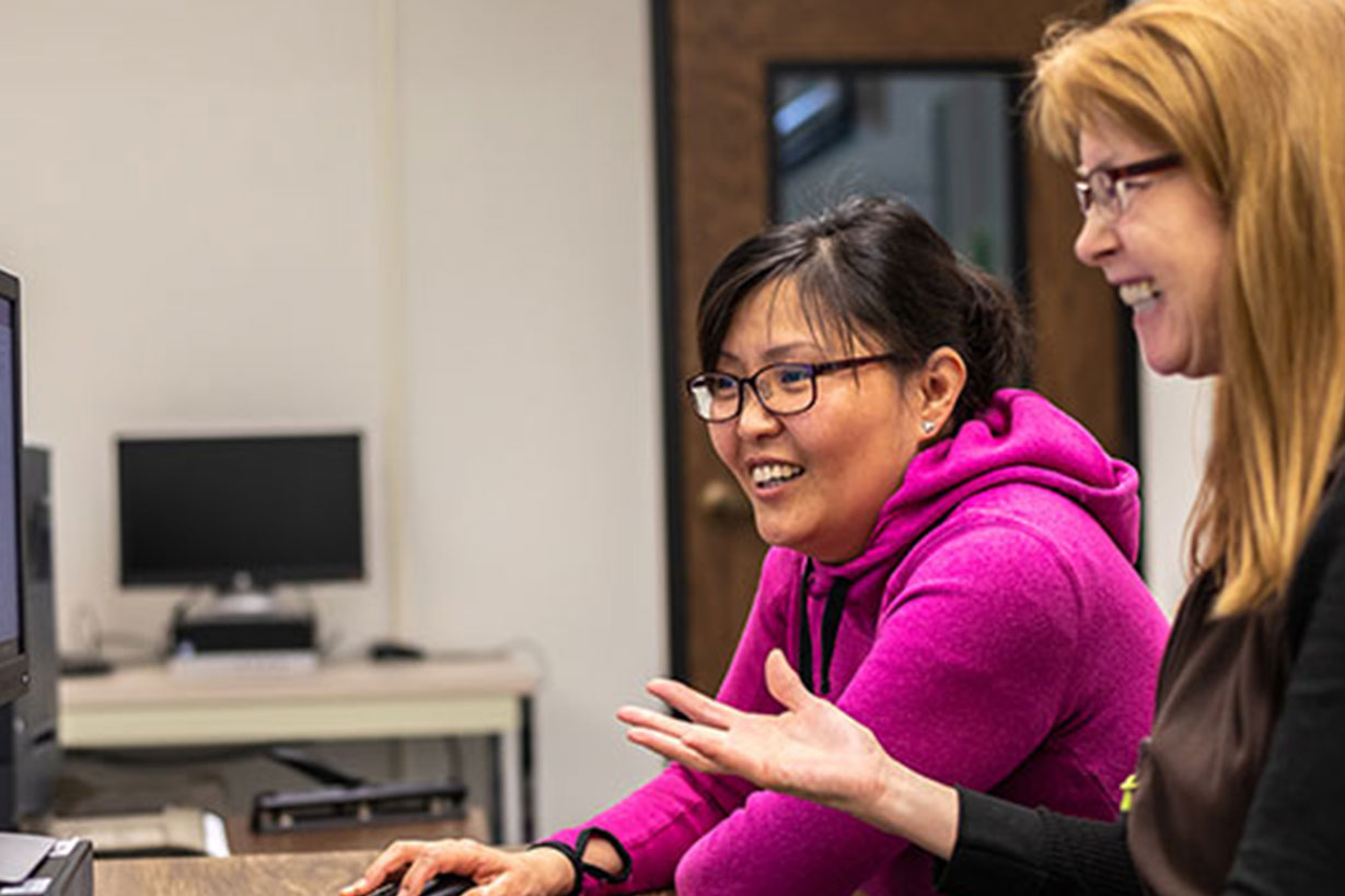 2 faculty members chat while sitting in front of a computer.
